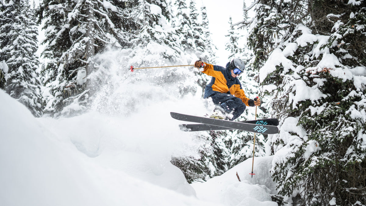 Skier in an orange and navy jacket airborne through deep powder between snow-loaded trees on Poet skis. Photo by Aaron Sheffner.